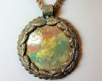 Puede incluir: Un collar de cadena dorada con un colgante redondo. El colgante está hecho de metal texturizado con una pátina verde y marrón. El centro del colgante es una pieza redonda e iridiscente de vidrio con un patrón amarillo, verde y naranja.