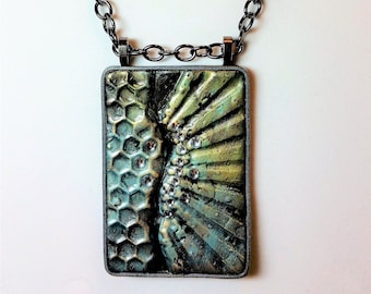 Puede incluir: Un collar de cadena plateada con un colgante rectangular. El colgante está hecho de resina verde y dorada con un patrón de panal en un lado y un patrón en forma de abanico en el otro. El colgante está decorado con pequeños diamantes de imitación transparentes.