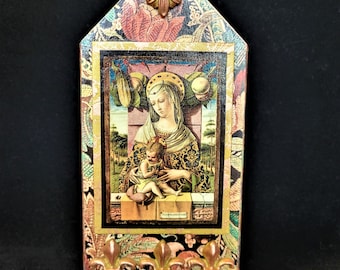 May include: A wooden plaque with a decorative floral design and a framed image of the Virgin Mary and baby Jesus. The plaque has a gold-colored metal fleur-de-lis design at the bottom.