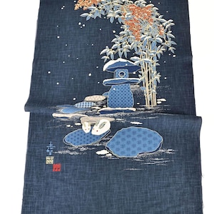 May include: Indigo blue fabric with a Japanese garden design. The illustration features a stone lantern, bamboo, and stylized rocks with rabbits. The design includes white accents and red berries, creating a serene, nature-inspired scene.