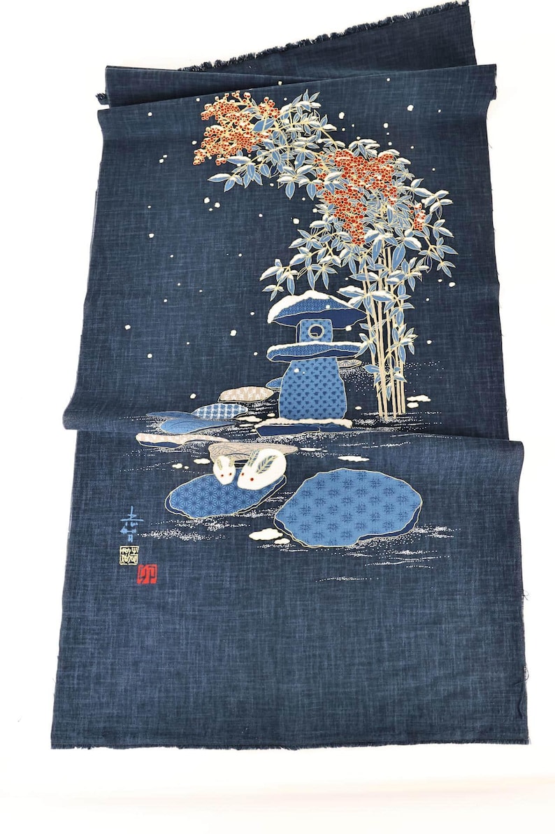 May include: Indigo blue fabric with a Japanese garden design. The illustration features a stone lantern, bamboo, and stylized rocks with rabbits. The design includes white accents and red berries, creating a serene, nature-inspired scene.
