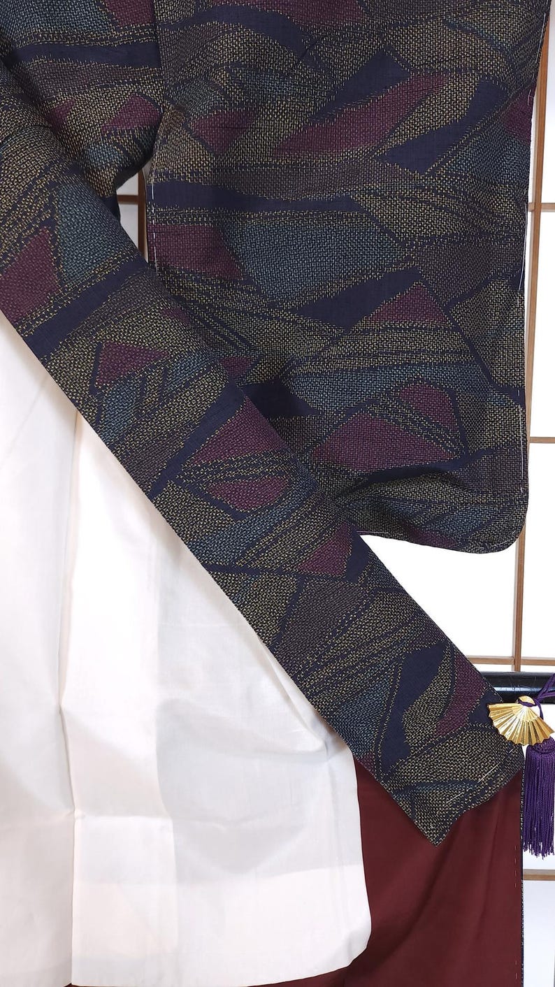 May include: Close-up of a patterned kimono with a geometric design in navy, burgundy, and gold. The kimono's interior is white, and the trim is a deep burgundy. A decorative tassel hangs from the side.