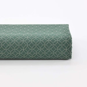 Peut inclure: Un tissu vert avec un motif de cercles et de lignes cousus en blanc. Le tissu est plié en deux et posé sur une surface blanche.