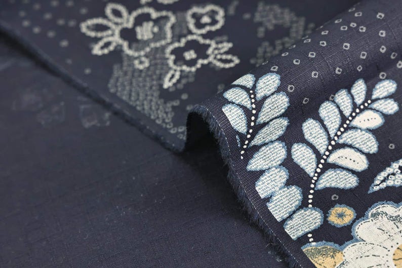 May include: Close-up of navy blue fabric with a floral and leaf pattern in white and light blue. The fabric is folded, revealing the detailed design and texture. The design includes stylized flowers and dotted accents.