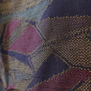 May include: Close-up of a woven fabric with a geometric pattern. The fabric features a dark blue background with diagonal stripes and diamond shapes in shades of gold, teal, and burgundy. The texture appears slightly textured.