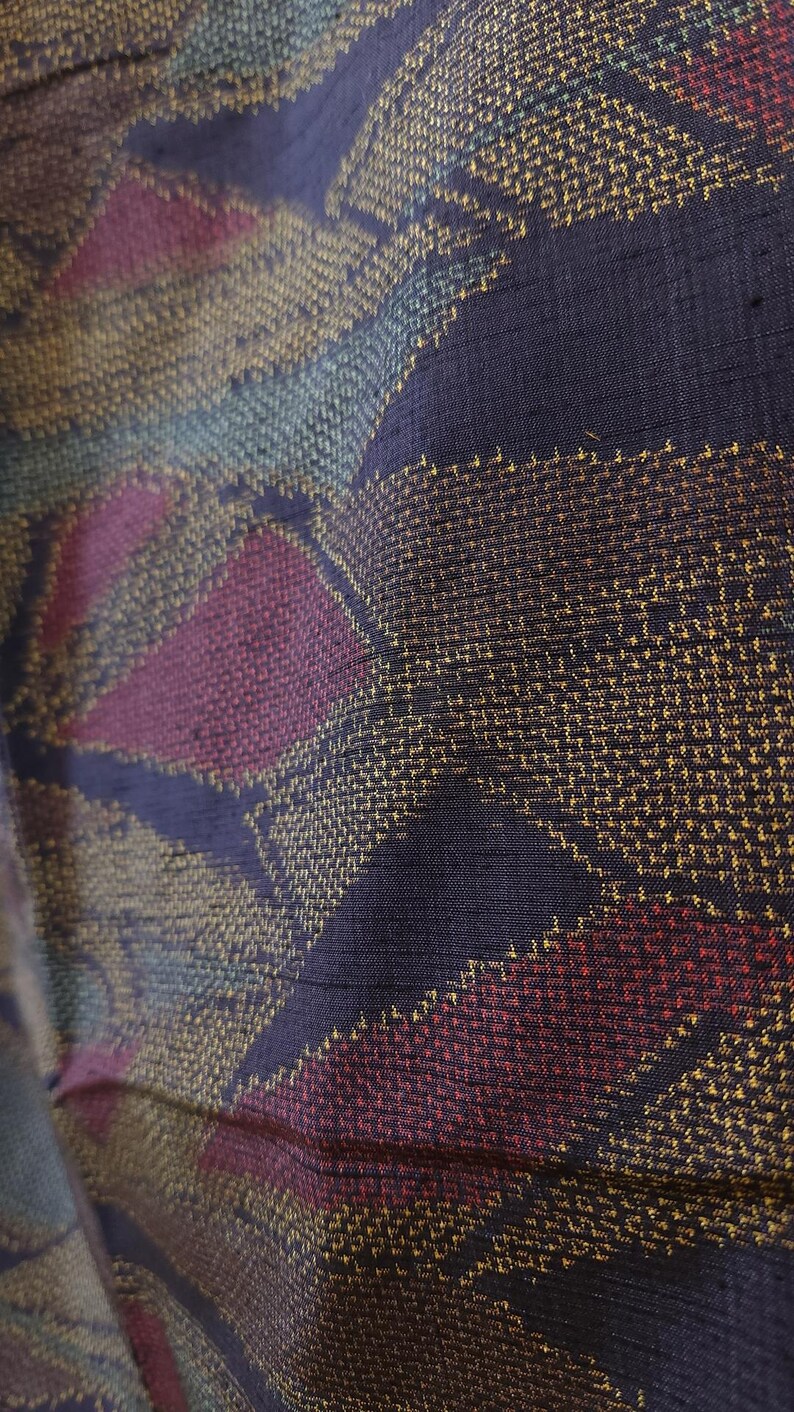 May include: Close-up of a woven fabric with a geometric pattern. The fabric features a dark blue background with diagonal stripes and diamond shapes in shades of gold, teal, and burgundy. The texture appears slightly textured.