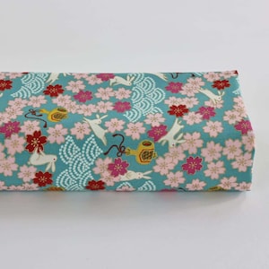 May include: A folded fabric with a Japanese-inspired design. The fabric features a teal background with white rabbits, pink and red cherry blossoms, and stylized wave patterns. Decorative elements include yellow and gold tea kettles.