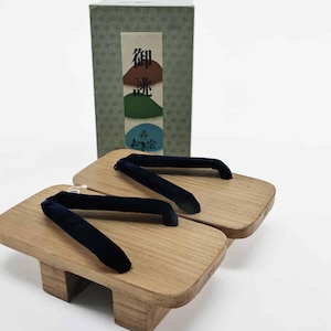 May include: A pair of traditional Japanese geta sandals with wooden platforms and black fabric straps. The sandals are displayed in front of a green box with Japanese text and colorful illustrations.