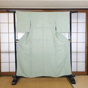 Peut inclure: Un kimono vert pâle est suspendu à un portant en métal noir. Le kimono a des manches longues et un design fluide. Le portant est placé devant des portes coulissantes japonaises traditionnelles et un tatami.