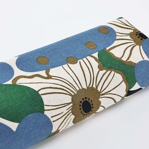 May include: Rolled fabric with a floral design. The pattern includes large white flowers with brown centres and green leaves, set against a blue background with blue and brown shapes. The fabric is likely cotton or linen.