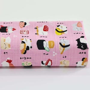 May include: Pink fabric featuring a repeating pattern of sushi and cat illustrations. The sushi and cats are in various colours, including black, white, yellow, and red. Japanese characters are also printed on the fabric.