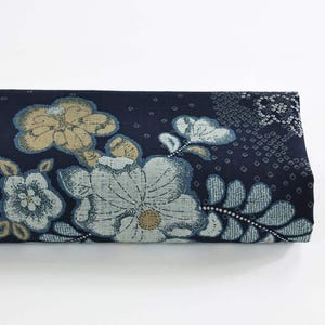 May include: Folded fabric with a navy blue background and a floral pattern. The flowers are in shades of light blue, beige, and white. The design includes detailed leaves and a dotted pattern.