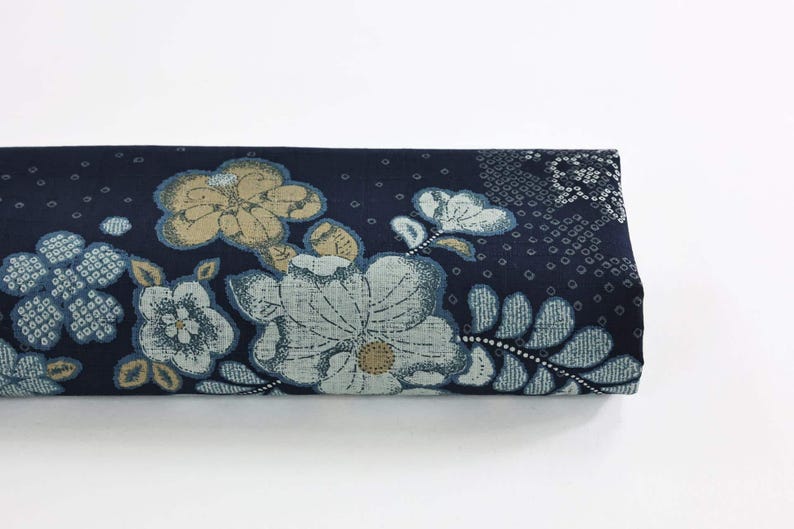 May include: Folded fabric with a navy blue background and a floral pattern. The flowers are in shades of light blue, beige, and white. The design includes detailed leaves and a dotted pattern.