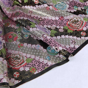 Può includere: Primo piano di un tessuto con motivo floreale. Il tessuto presenta colori rosa, nero e oro con dettagliati disegni floreali e geometrici. I bordi sono rifiniti con un bordo nero.