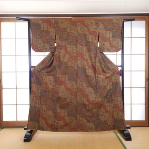 May include: A brown kimono featuring a repeating pattern of diagonal red and blue wave-like motifs overlaid with thin vertical and horizontal black lines. The garment is displayed on a black wooden stand with purple tassels at the sides.
