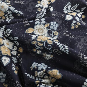 May include: Close-up of a dark blue fabric with a floral pattern. The design features white, light blue, and gold flowers and leaves. The fabric is folded, showing the texture and detail of the print.