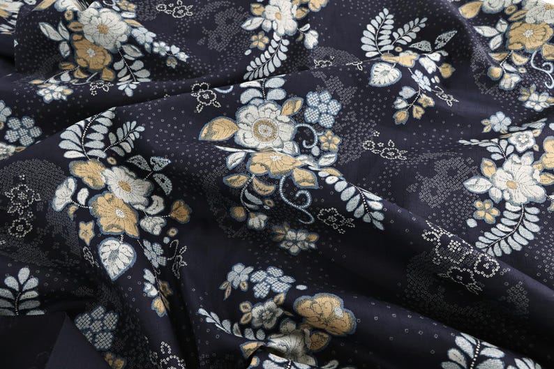May include: Close-up of a dark blue fabric with a floral pattern. The design features white, light blue, and gold flowers and leaves. The fabric is folded, showing the texture and detail of the print.