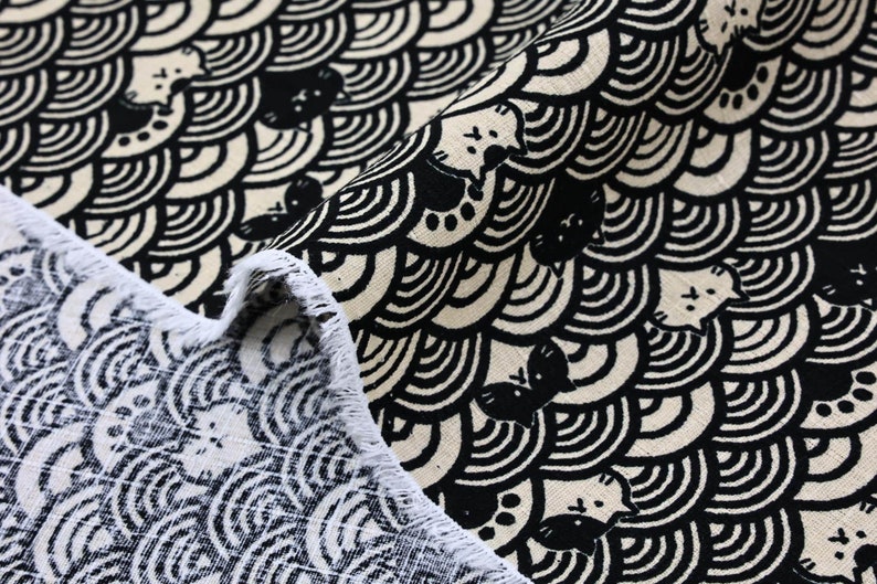 Op de afbeelding: Zwart-witte stof met een herhalend patroon van gestileerde katten en golven. De stof heeft een rafelige rand.
