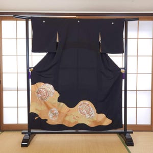 May include: A black kimono with a decorative floral pattern in shades of gold, peach, and white. The kimono is displayed on a black stand in front of a traditional Japanese sliding door.