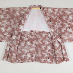 May include: A kimono with a brown and white speckled pattern. The garment is open, revealing a white and pink interior. The kimono is hanging from a wooden hanger against a plain white background. The sleeves are wide and the fabric appears lightweight.