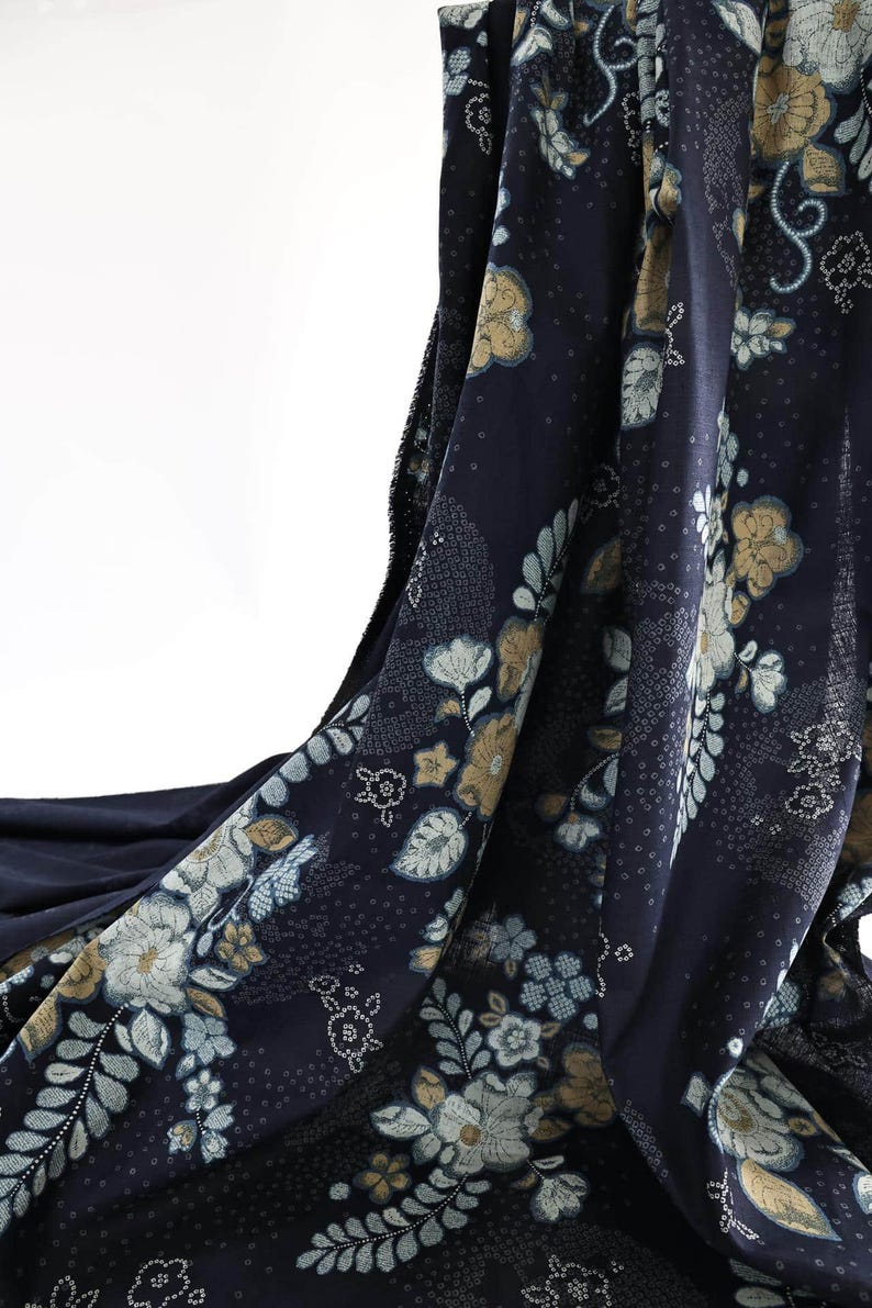 May include: Navy fabric with a floral design in white, pale blue, and gold. The fabric is draped, revealing the intricate pattern of flowers and leaves. The background is white.