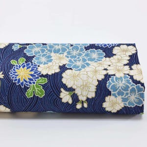 Peut inclure: Un tissu rectangulaire plié, avec un fond bleu foncé orné d'un motif floral blanc, bleu clair et jaune. Le motif comprend des fleurs stylisées et des motifs en forme de vagues.