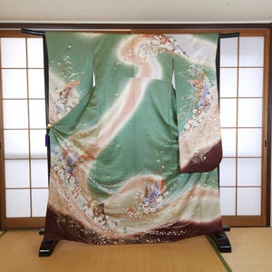 Peut inclure: Un kimono vert avec un motif floral, présenté sur un support noir. Le kimono présente un motif en forme de vagues dans des tons de rose et de blanc, avec des motifs floraux et d'oiseaux détaillés. Les manches ont une bordure marron foncé.
