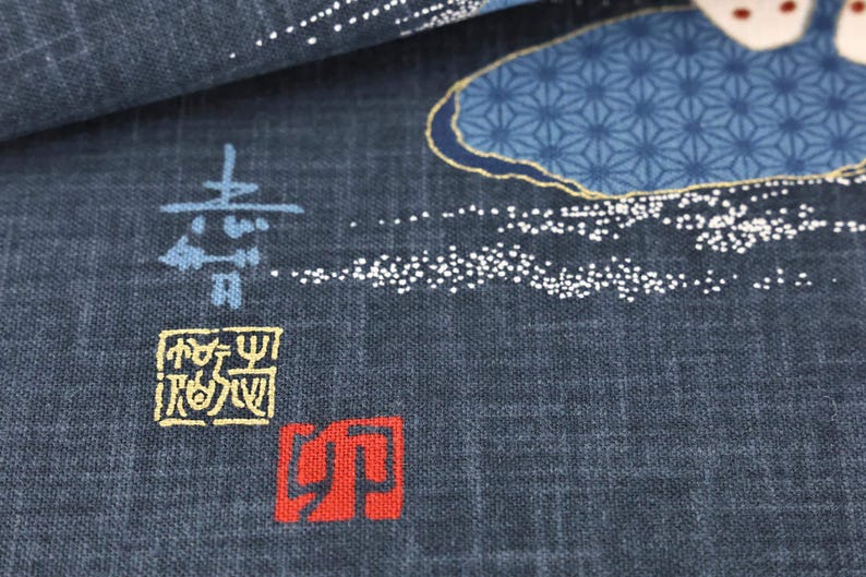 May include: Close-up of a dark blue fabric with Japanese-style designs. The fabric features blue and gold floral patterns, white dotted accents, and Japanese characters in blue, yellow, and red.