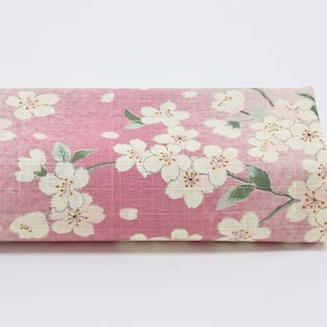 Peut inclure: Un rouleau de tissu plié avec un fond rose orné d'un motif de fleurs de cerisier blanches et de feuilles vertes. Le tissu a un aspect texturé, peut-être du lin. Le motif évoque le printemps et la nature, avec un motif floral délicat.