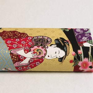 May include: A close-up of a fabric with a Japanese-inspired design. The fabric features a woman in a red kimono holding a fan, surrounded by gold and blue geometric patterns and pink flowers.