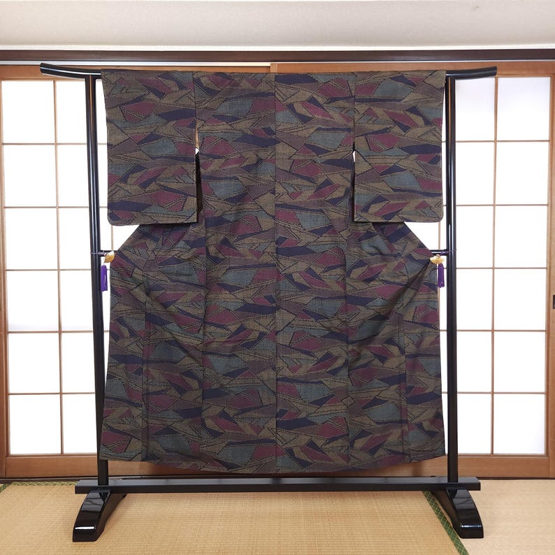 May include: A patterned kimono in shades of brown, burgundy, and gray is displayed on a black metal rack. The fabric features an abstract geometric design. The kimono is hanging in front of a traditional Japanese sliding door.