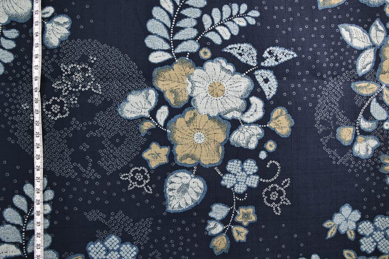 May include: Dark navy blue fabric with a floral pattern. The design features large flowers in shades of white, tan, and light blue, along with smaller floral and dot patterns. A measuring tape is visible along the left side.