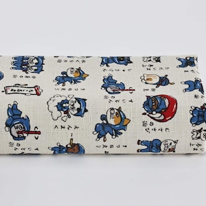 Op de afbeelding: Gevouwen stof met een herhalend patroon van cartoon ninja katten in blauw, wit en rood. De katten zijn in verschillende actieposities, met Japanse tekst. De stof heeft een lichtbeige achtergrond. Afmetingen in centimeters.