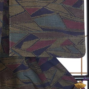 May include: Close-up of a patterned textile with a geometric design in shades of navy, burgundy, teal, and gold. The fabric appears to be woven, with a textured surface. The textile is likely part of a garment.