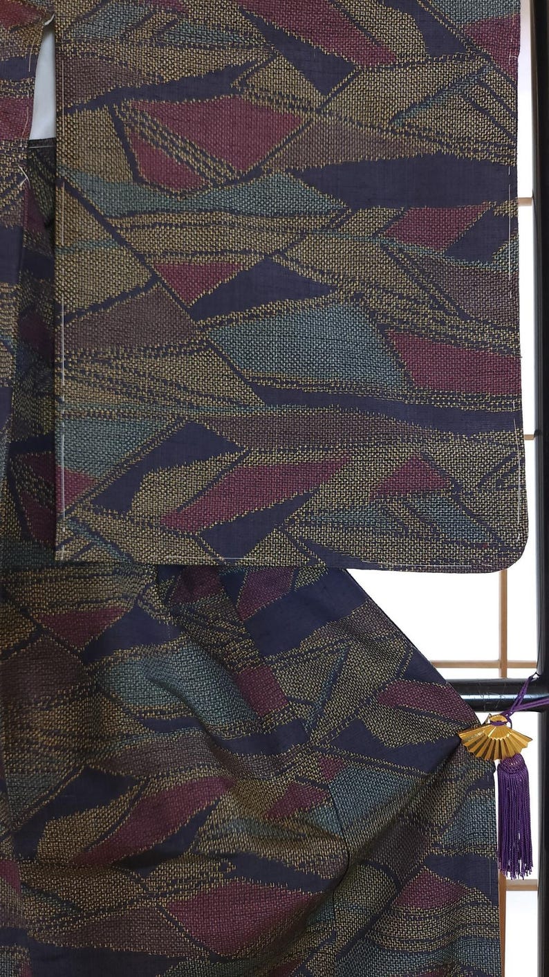 May include: Close-up of a patterned textile with a geometric design in shades of navy, burgundy, teal, and gold. The fabric appears to be woven, with a textured surface. The textile is likely part of a garment.