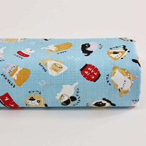 May include: A rolled-up piece of light blue fabric with a repeating pattern of cartoon cats, food items, and Japanese text. The cats are in various poses and outfits, with some wearing hats or holding food. The fabric has a textured appearance.