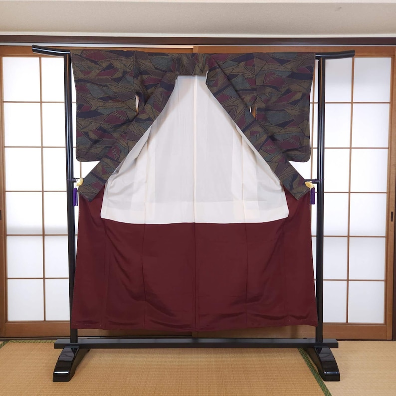 May include: A traditional Japanese kimono displayed on a black stand. The kimono features a patterned upper section in shades of green, purple, and brown, with a white inner lining and a deep red lower section. The garment is presented against a backdrop of sliding doors.
