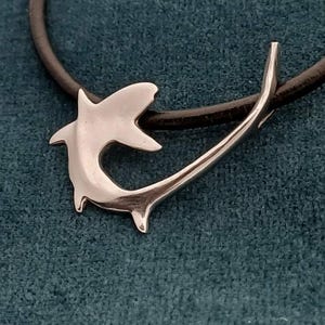 May include: A silver-colored shark pendant necklace with a black leather cord. The shark pendant is stylized with a curved body and a pointed fin. The necklace is displayed on a teal-colored surface.