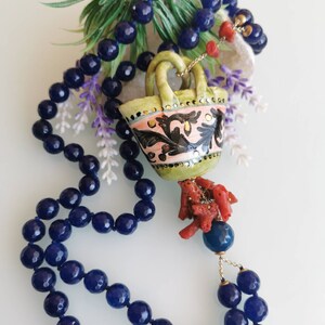 May include: A long necklace made of dark blue beads with a ceramic pendant shaped like a basket with a coral tassel. The basket is decorated with pink, green, and gold details.