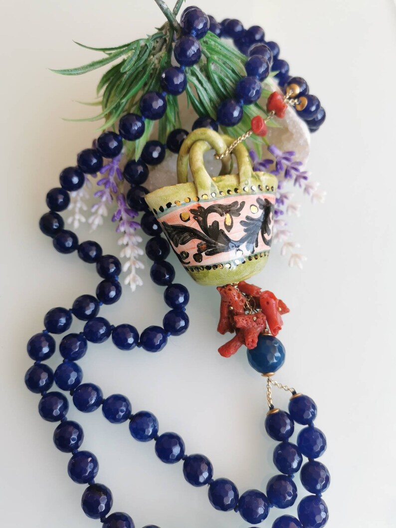 May include: A long necklace made of dark blue beads with a ceramic pendant shaped like a basket with a coral tassel. The basket is decorated with pink, green, and gold details.
