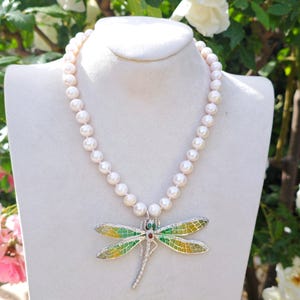 Puede incluir: Un collar de perlas blancas con un colgante de libélula de plata. La libélula tiene alas de esmalte verde y amarillo con acentos rojos.