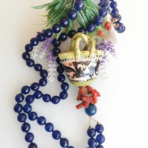 May include: A blue gemstone necklace with a ceramic basket pendant and coral accents. The basket is decorated with floral patterns in pink, green, and blue.
