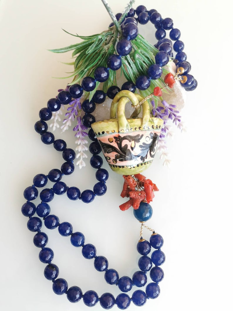 May include: A blue gemstone necklace with a ceramic basket pendant and coral accents. The basket is decorated with floral patterns in pink, green, and blue.