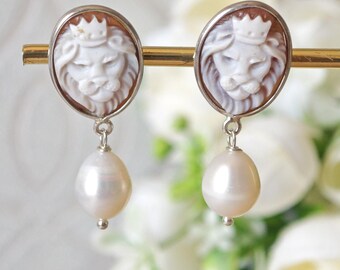 May include: A pair of drop earrings featuring oval-shaped cameos with a carved white lion wearing a crown against a brown background, set in a silver-toned metal frame. Each earring includes a dangling, teardrop-shaped white pearl at the base.