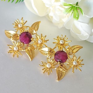 Puede incluir: Pendientes de tono dorado con un diseño floral con piedras preciosas rosas y detalles de perlas blancas.