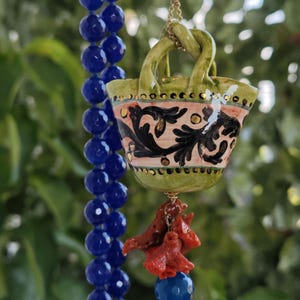 May include: A green ceramic basket with a pink, black, and gold floral design hangs from a blue beaded necklace with coral accents.