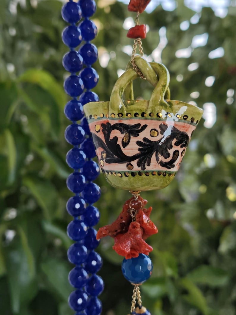 May include: A green ceramic basket with a pink, black, and gold floral design hangs from a blue beaded necklace with coral accents.