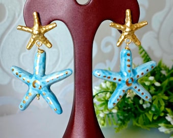 Könnte beinhalten: Ein Paar Ohrringe mit Seestern-Design. Die Ohrringe haben einen goldfarbenen Seestern-Aufsatz und einen hellblauen Seestern-Anhänger mit goldenen Akzenten. Die Ohrringe werden auf einem dunkelroten Ohrringständer präsentiert.