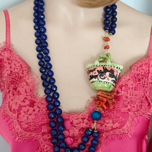 May include: A blue beaded necklace with a ceramic pendant featuring a colorful floral design. The pendant is attached to a coral and gold chain.