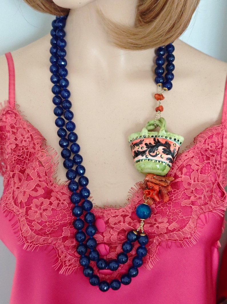 May include: A blue beaded necklace with a ceramic pendant featuring a colorful floral design. The pendant is attached to a coral and gold chain.